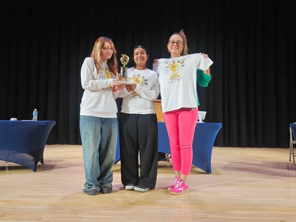 Two young ladies are wearing matching shirts and holding a trophy while an adult stands next to them and is holding a matching shirt.