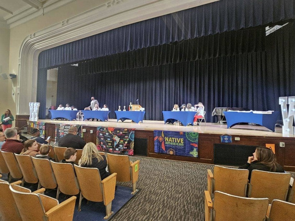 Multicultural Quiz Bowl contestants on stage at their tables while audience watches in auditorium.
