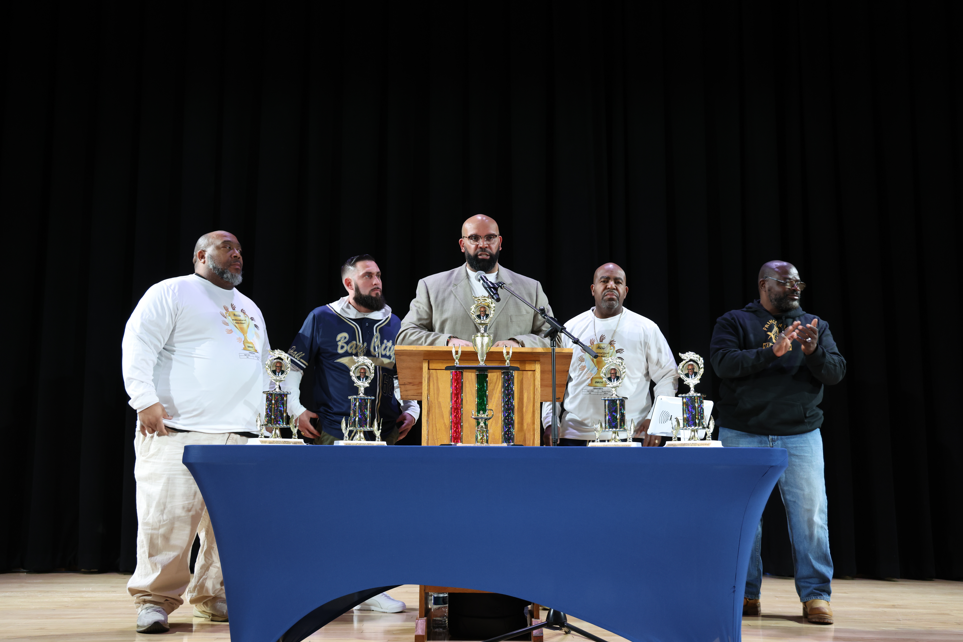 Individuals standing at a podium and there is a table with trophies in front of him. 