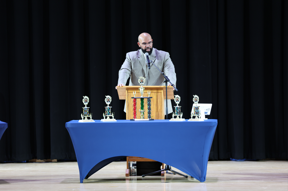 A man speaking at a podium and there is a table with trophies in front of him. 