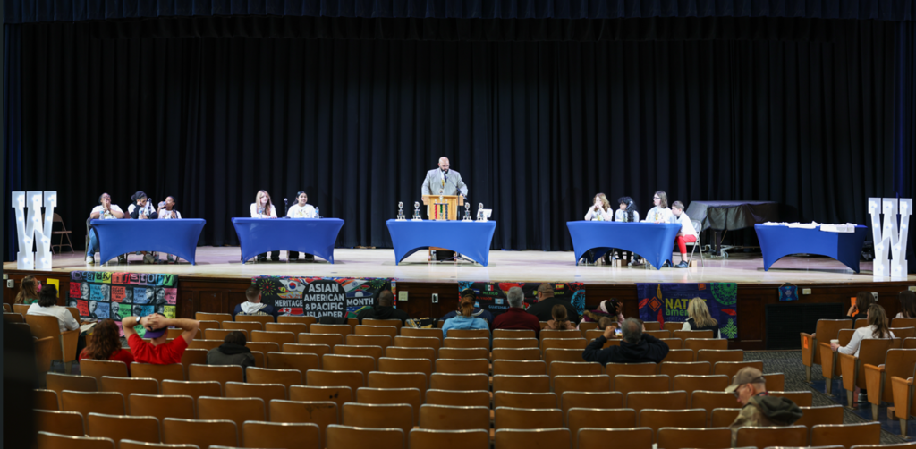 Students sitting behind a table and are competing in the multicultural quiz bowl. 