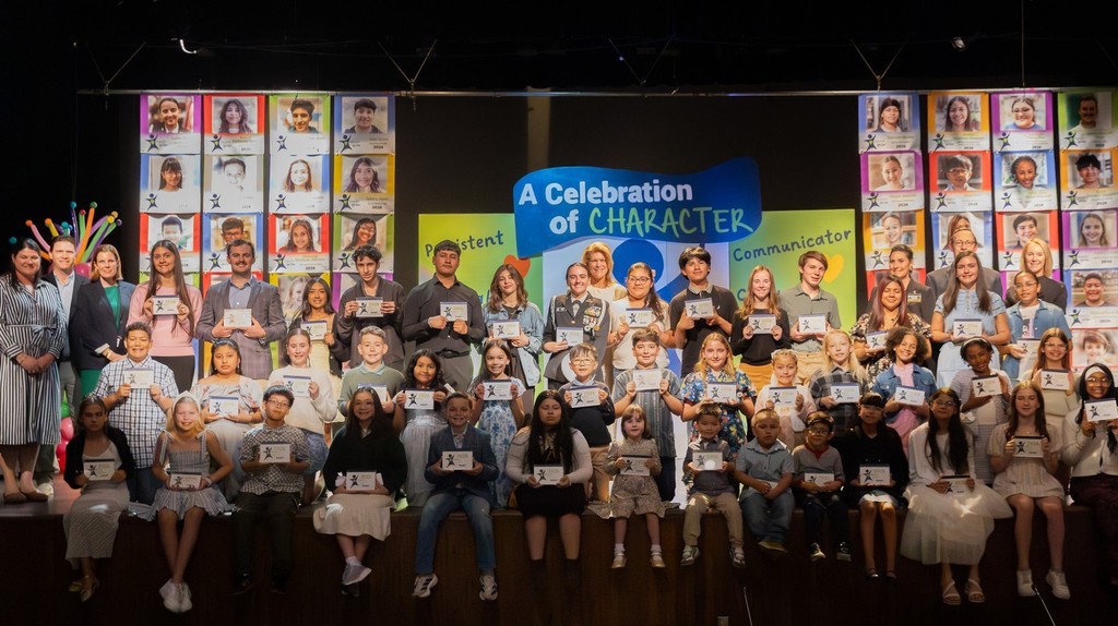 A group of students and adults stand on a stage holding certificates. They are in front of a colorful backdrop.