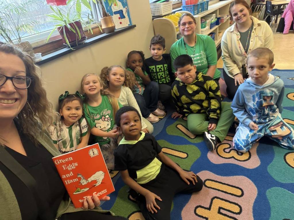Teacher and students wearing green while the teacher hold the "Green Eggs and Ham" book by Dr. Seuss.