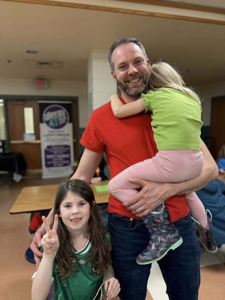A man holding a child in one and is smiling next to another child who is holding up to fingers and smiling. 