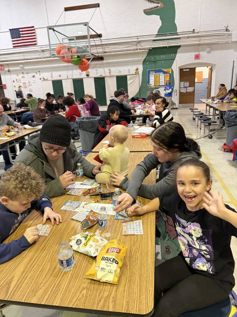 Parents and students writing, eating snacks, and talking in a school cafeteria.