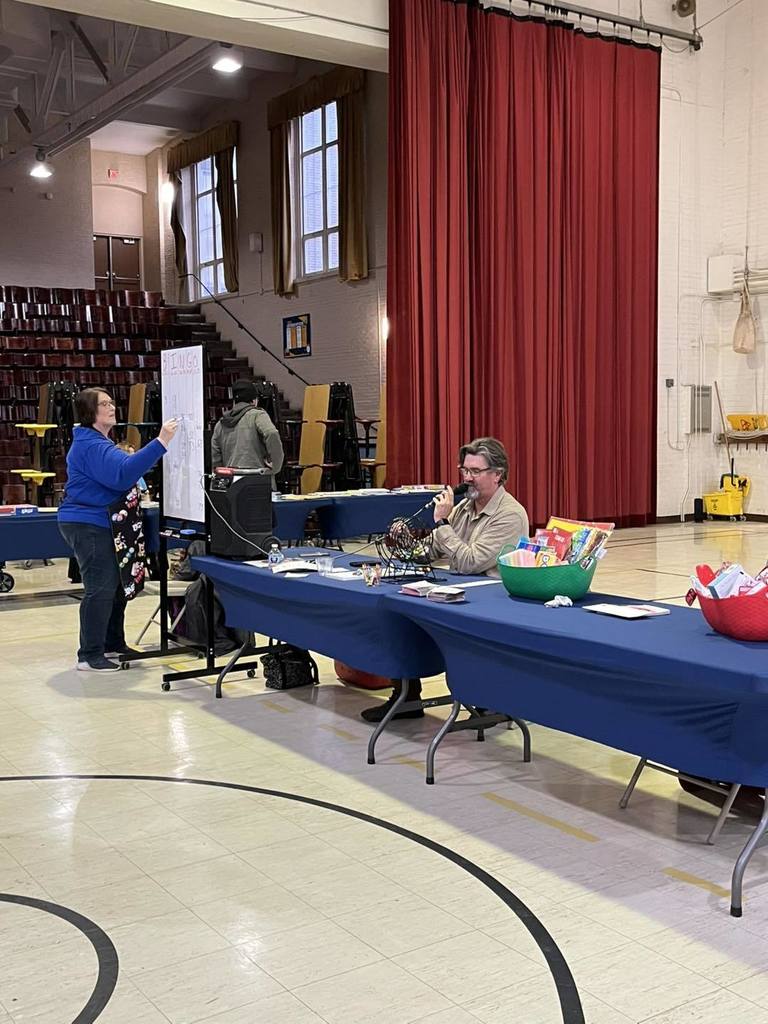 A man announcing numbers from a long table while a women writes on a white board.
