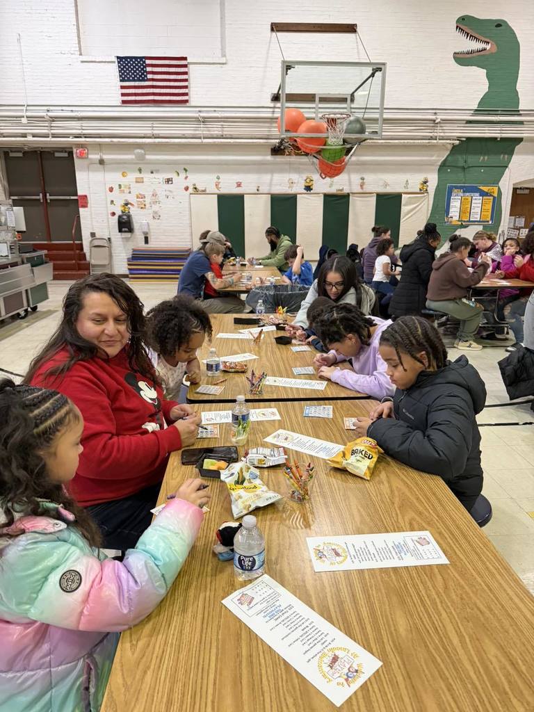 Parents and students writing, eating snacks, and talking in a school cafeteria.