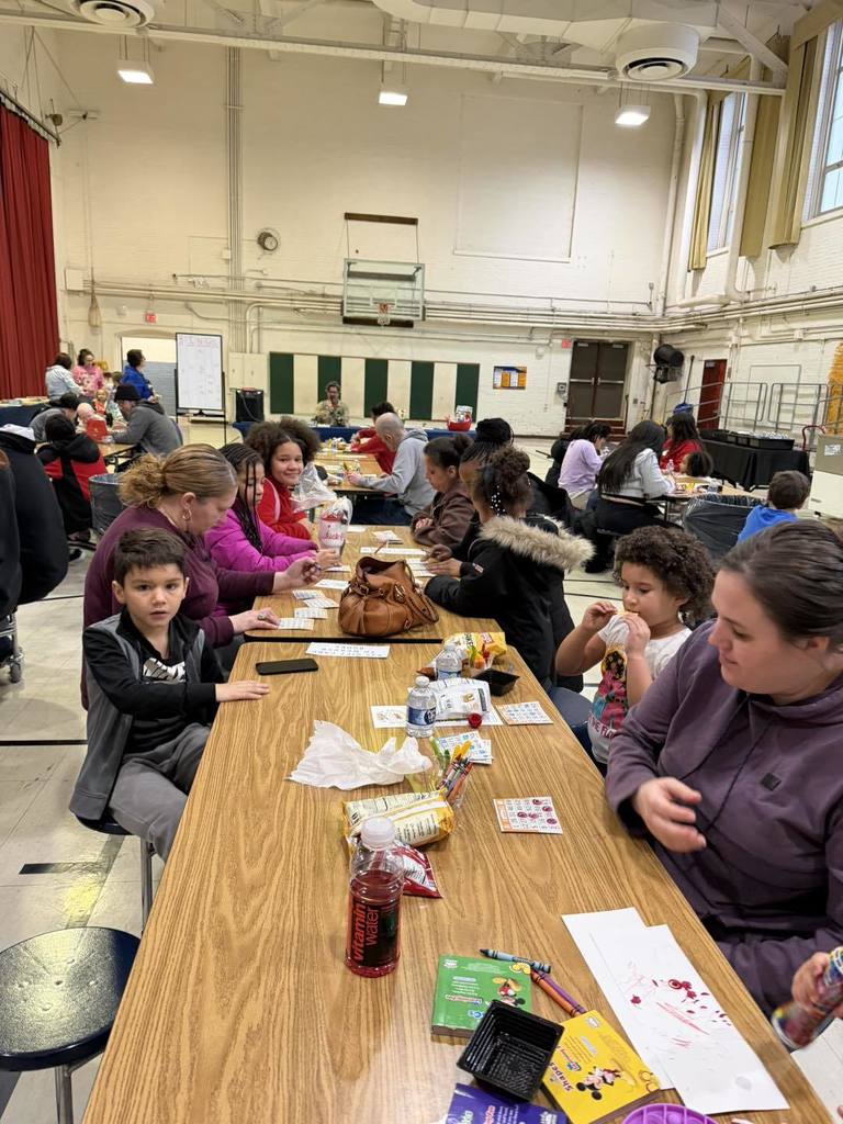 Parents and students writing, eating snacks, and talking in a school cafeteria.