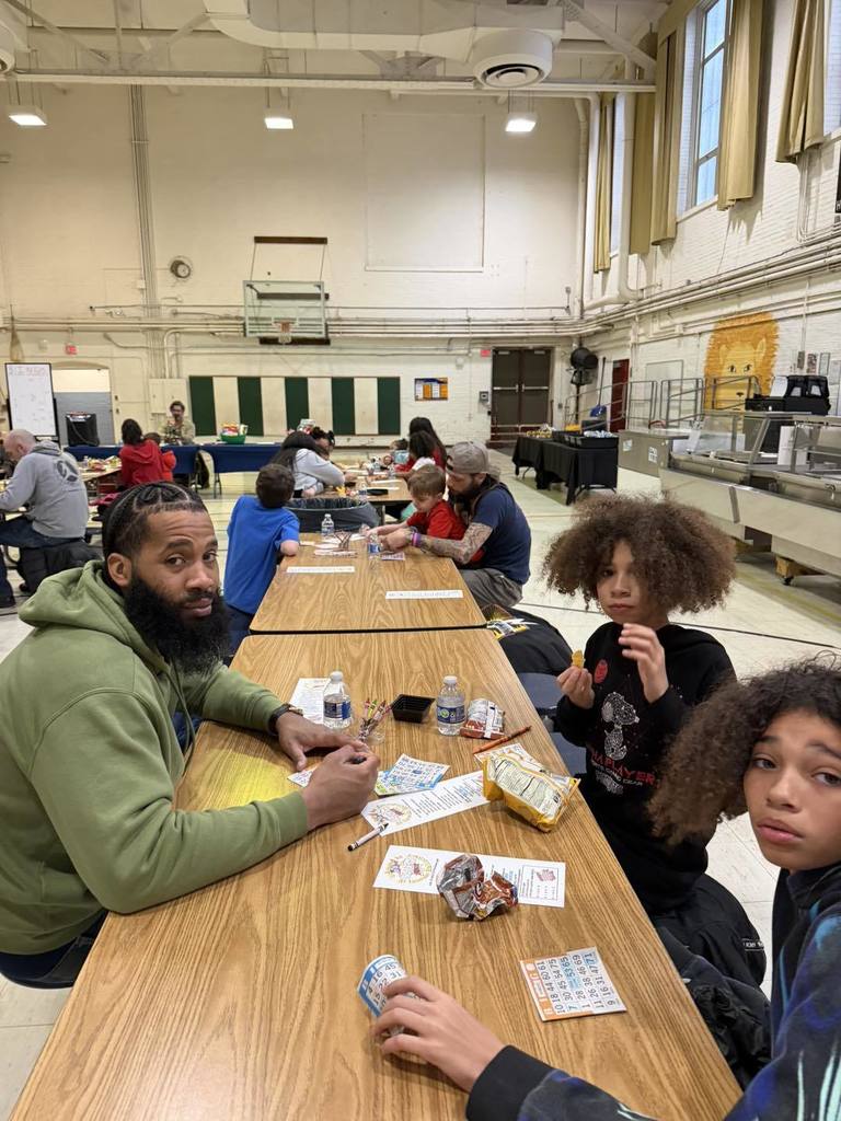 Parents and students writing, eating snacks, and talking in a school cafeteria.