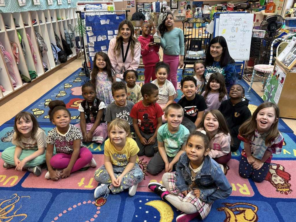 Students and teacher smiling in a classroom while one student holds a trophy. 