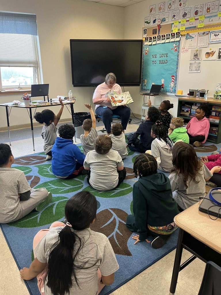 A women reading a book to a classroom of students. 