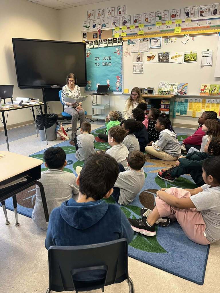 A women reading a book to a classroom of students. 