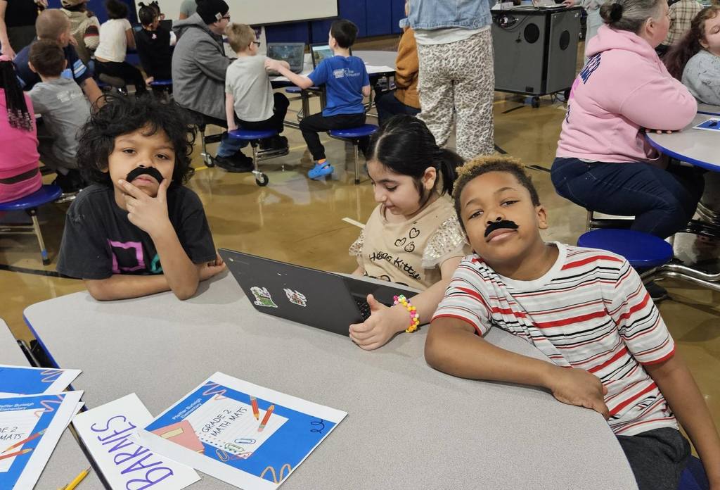 Students wearing fake mustaches and doing work at a table in the cafeteria. 