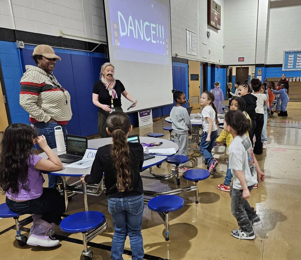 Teachers and students wearing fake mustaches and doing work at a table in the cafeteria. 