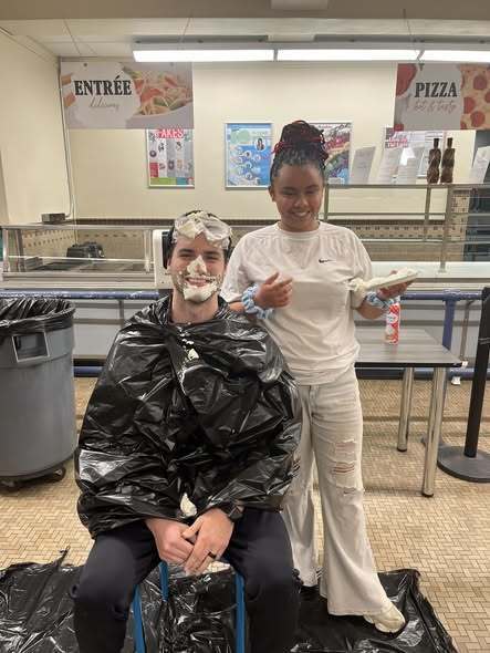 A student had pied a teacher in the school cafeteria and the teacher is wearing a black trash bag and smiling.