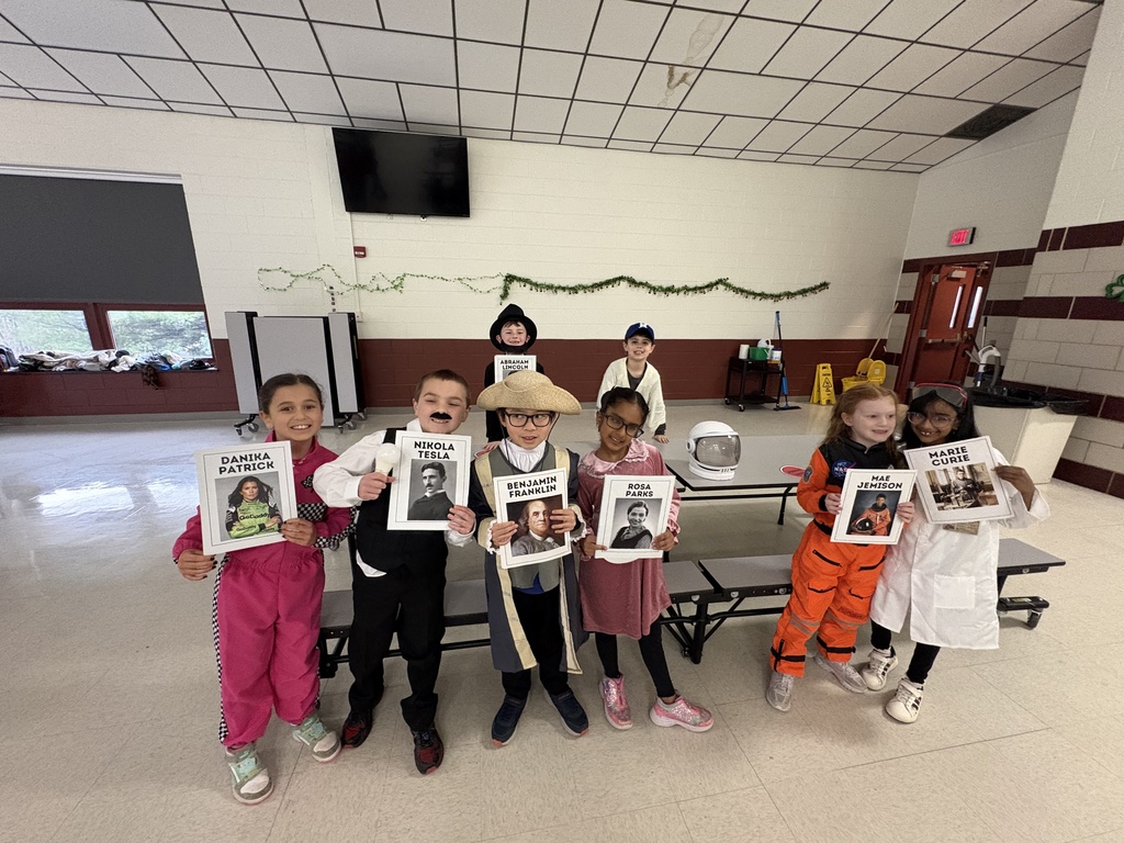 Students smiling with their posters. Famous figures include Danika Patrick, Nikola Tesla, Ben Franklin, Rosa Parks, Mar Jemison, Marie Curie, Abraham Lincoln.