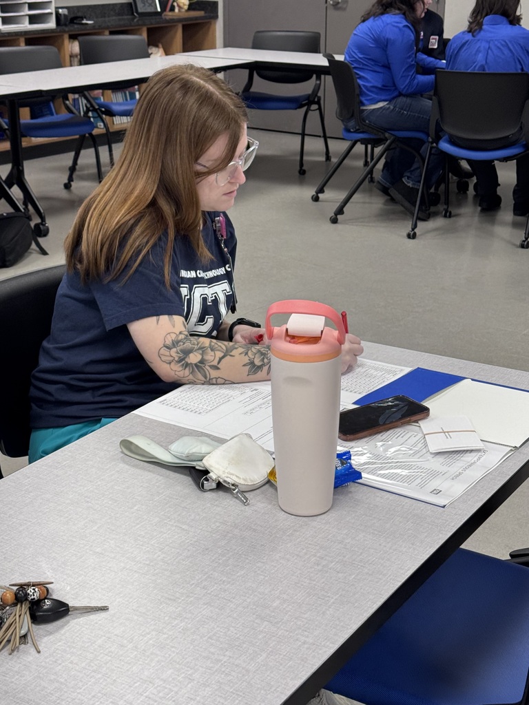 A woman sits at a desk, focusing on documents. A tumbler and phone rest on the table. Other people are seated nearby.