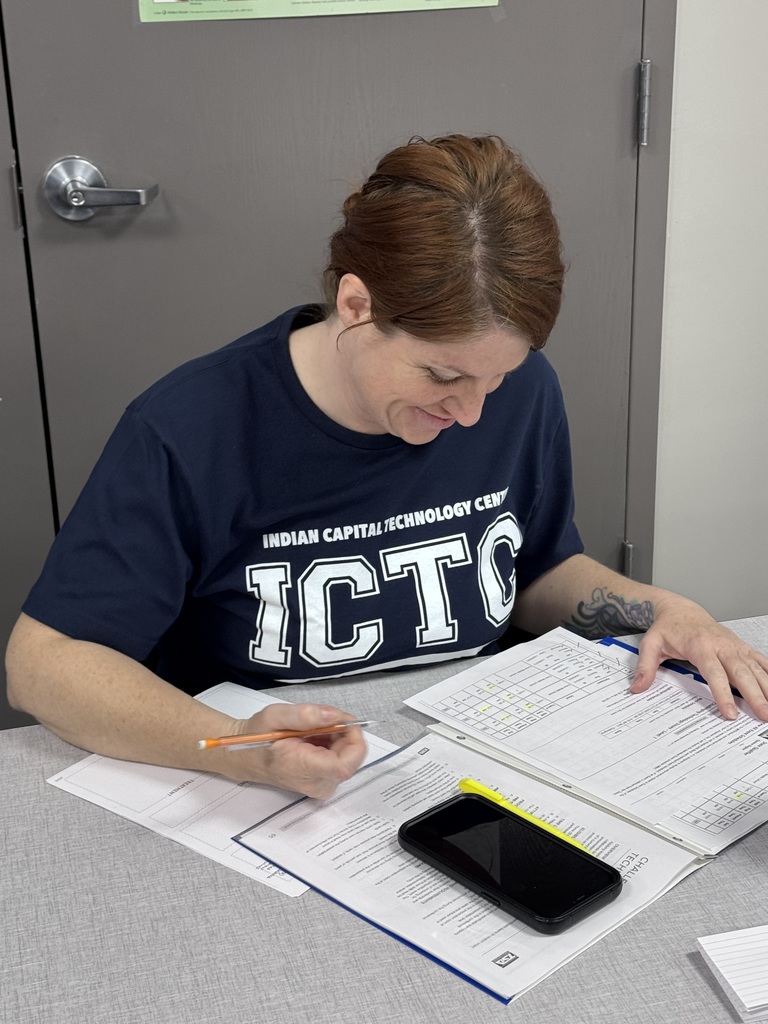 Person with short hair wearing a navy blue shirt with white letters. They are seated at a table with papers and a smartphone.