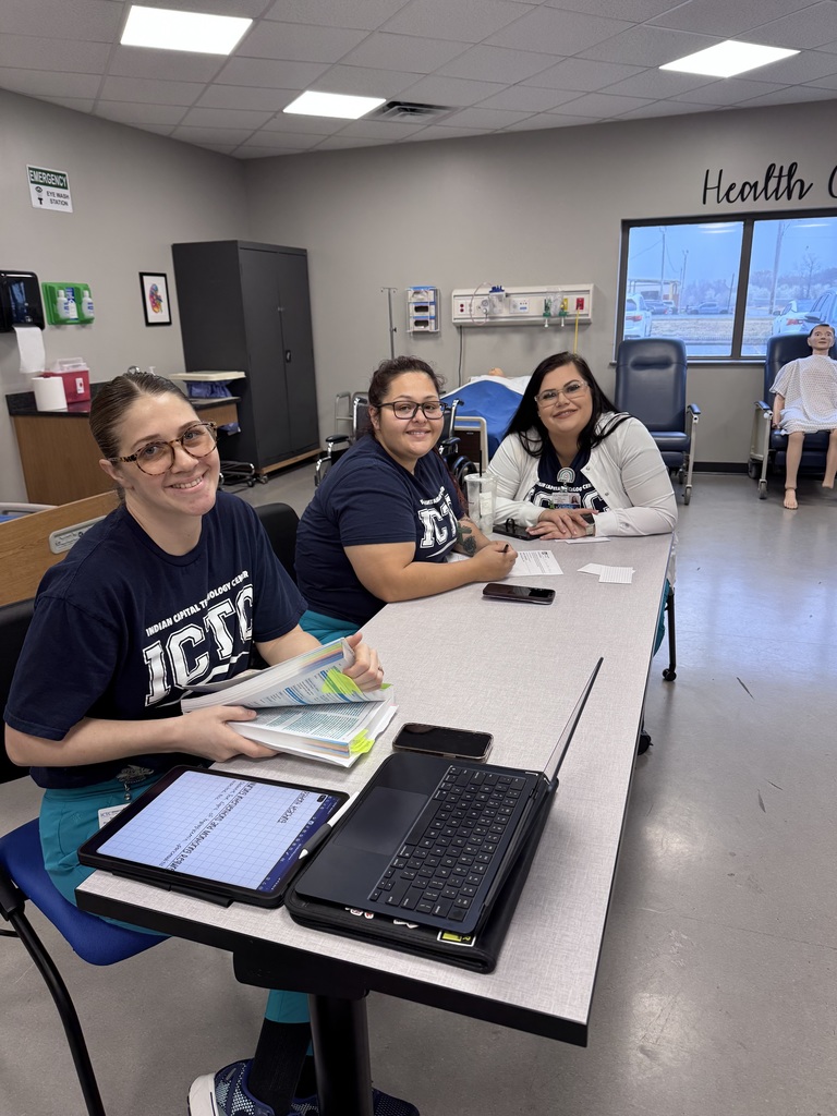 Three women seated around a table with a laptop, tablet, and book. Behind them, a room with medical equipment and a window.