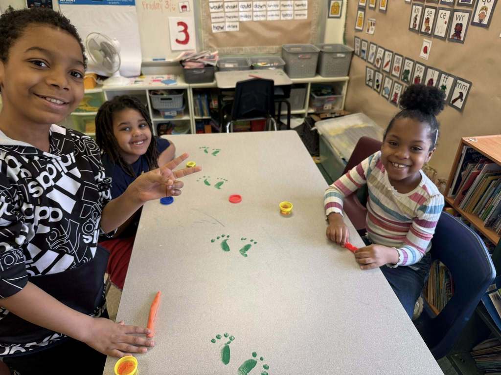 Kids playing with playdough on a table that has green foot prints on it. 