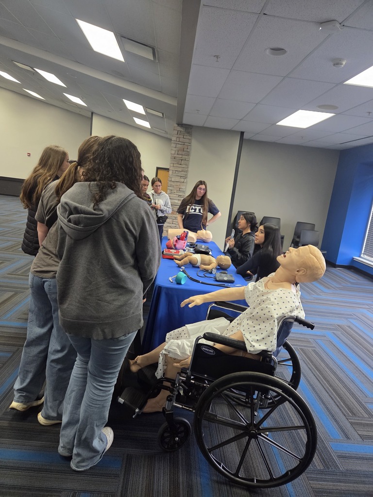 Several people gather around a table with a mannequin in a wheelchair. A woman in a hoodie stands nearby.