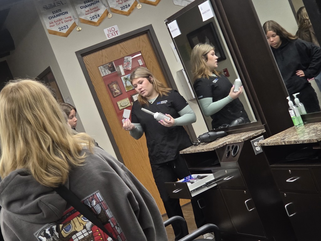 Four women in black uniforms in a salon, one holding a brush, others watching, and a mirror reflecting them.