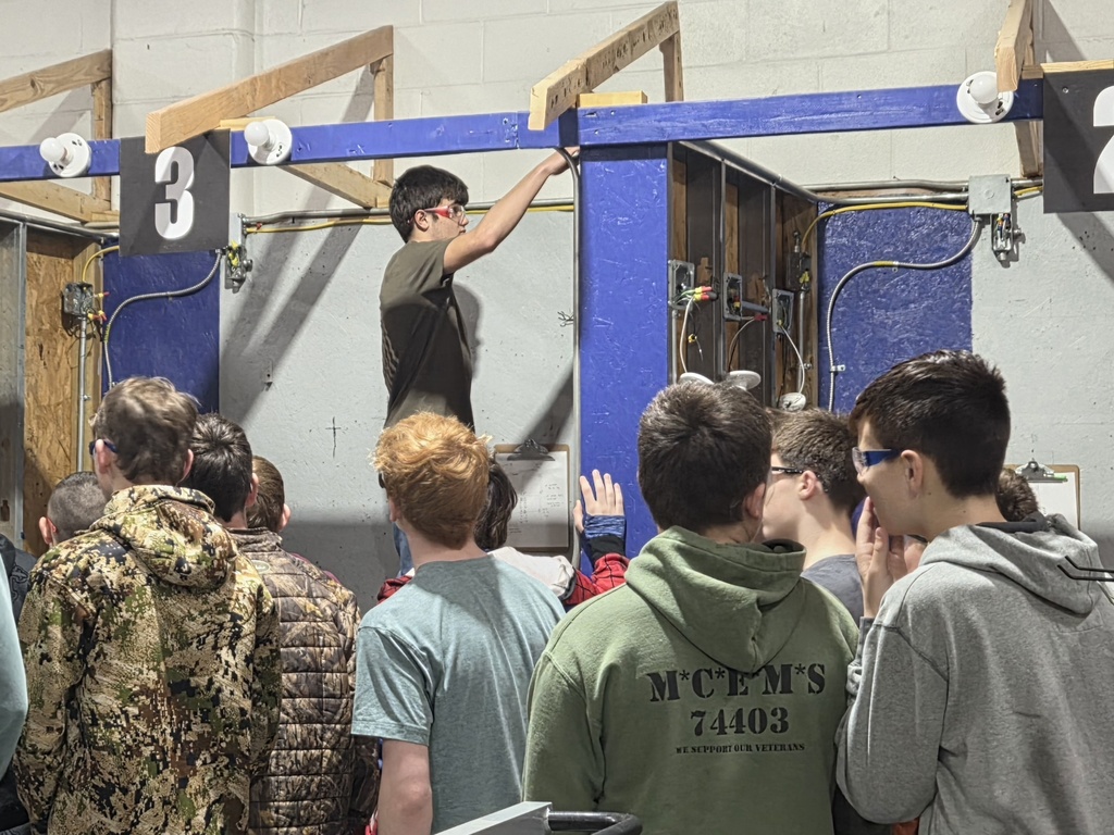 A group of students watch a man in a green shirt standing behind a blue box with the number 3 on it.