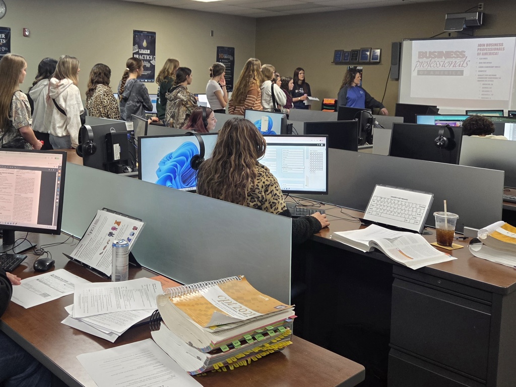 People sit at computers in an office. Some are seated at desks with papers and monitors. Others stand near a projector screen.