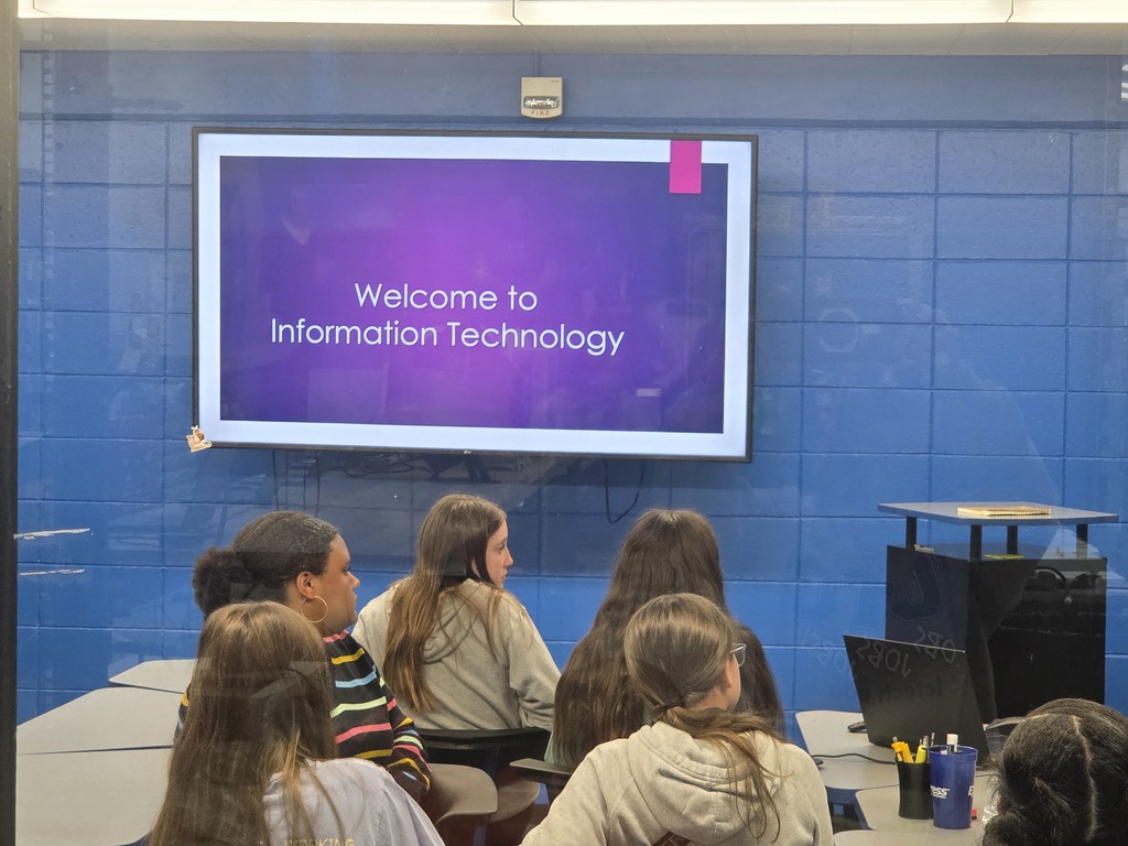 A classroom with blue walls. A screen displays "Welcome to Information Technology." Students are seated, facing the screen.