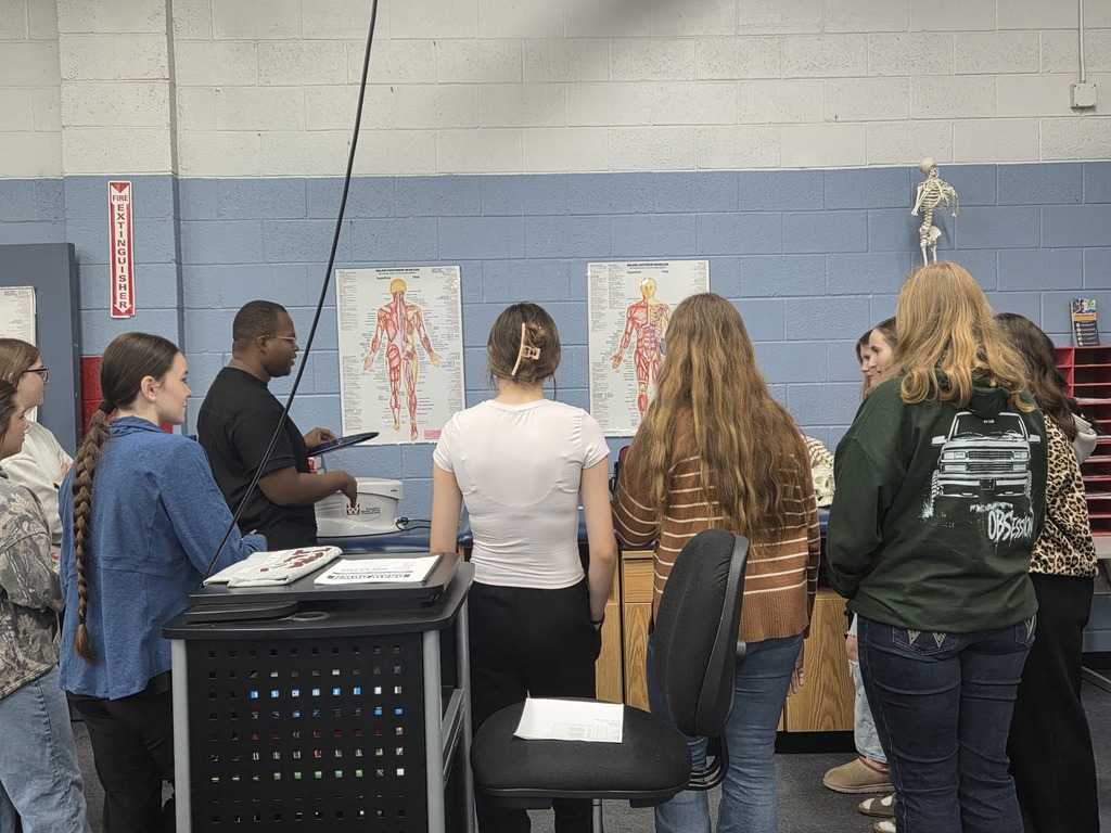 A group of people stand in a room with blue walls. A man stands at a desk, facing them.