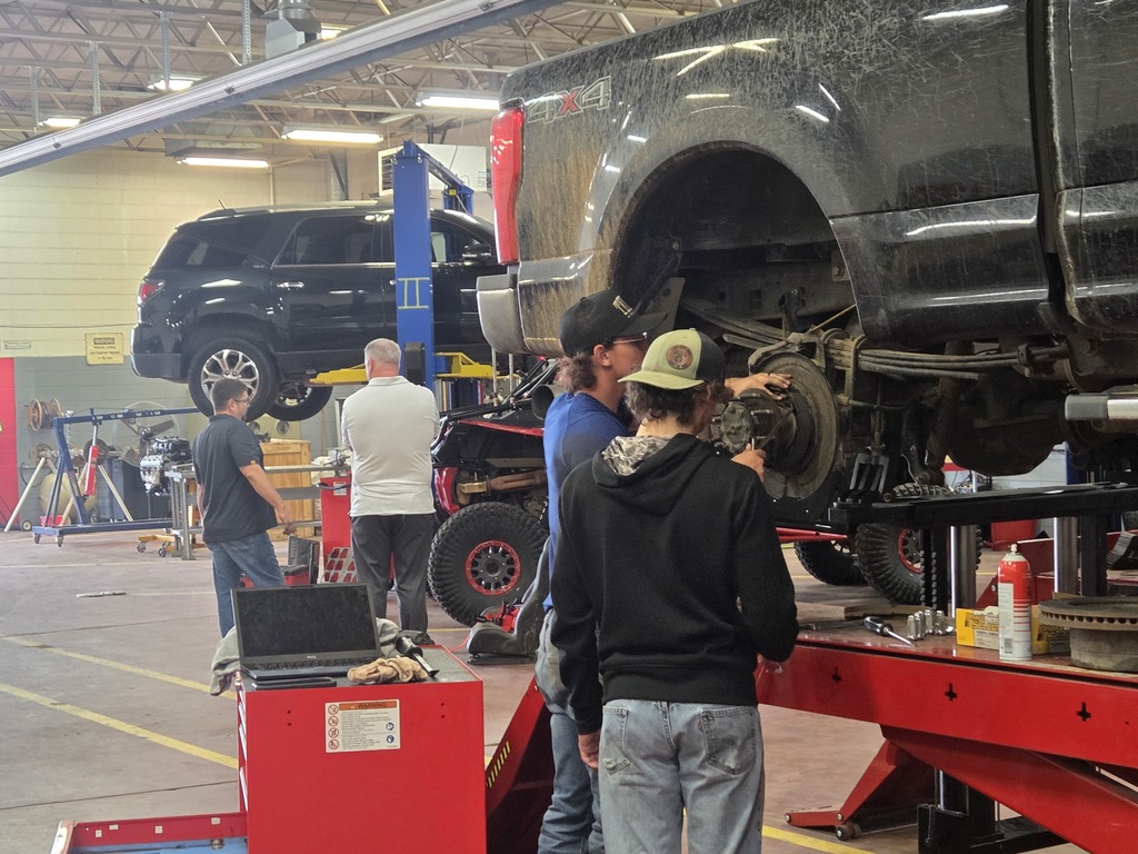 People in a workshop. A car is lifted on a jack, and four people are standing around it.