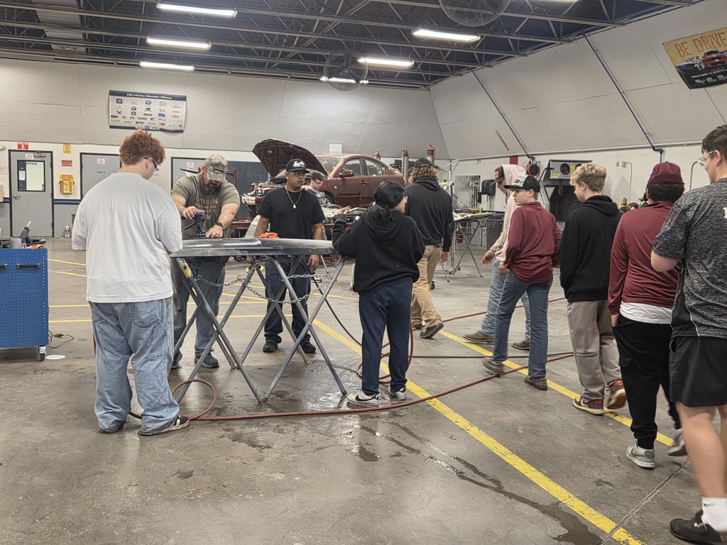 A group of people in a large room working on a car. Some are wearing jackets.