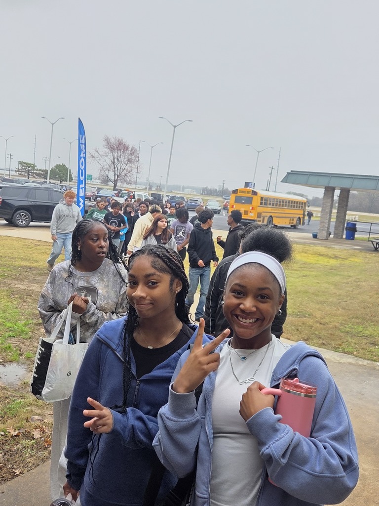 Group of people outside, one woman wearing a headband poses with a finger sign. Background includes a school bus and grassy area.