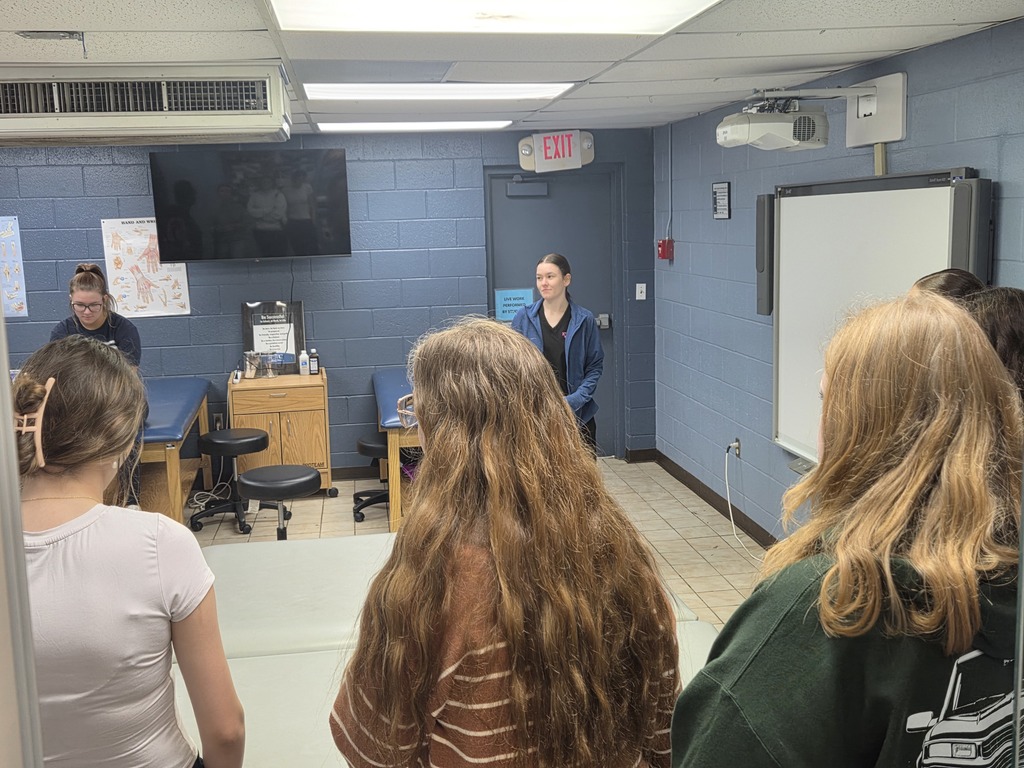 People in a classroom with a teacher at the front. A whiteboard and a television are mounted on the wall.