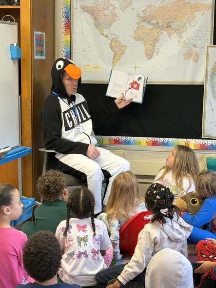 An adult dressed as a penguin and is reading a book to students in a classroom.