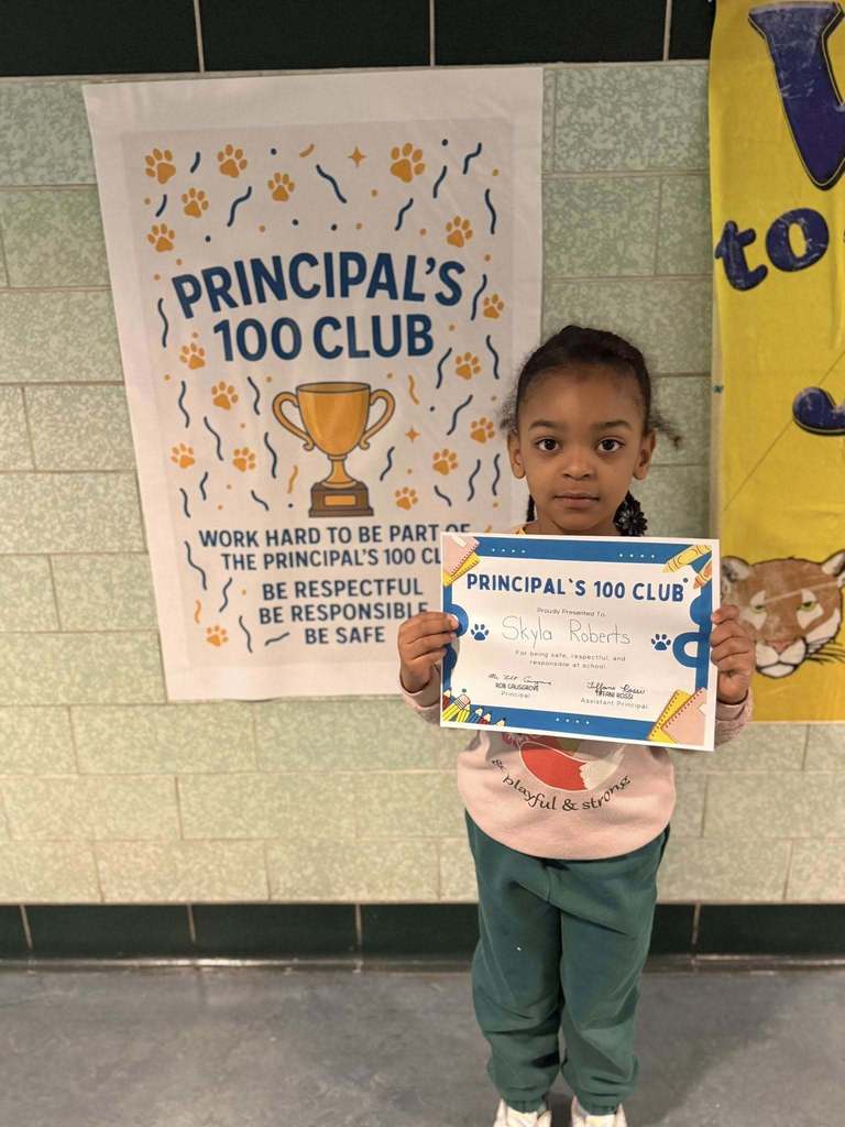 A young girl that is wearing a pink shirt and green pants is holding his Principal's 100 Club certificate. 