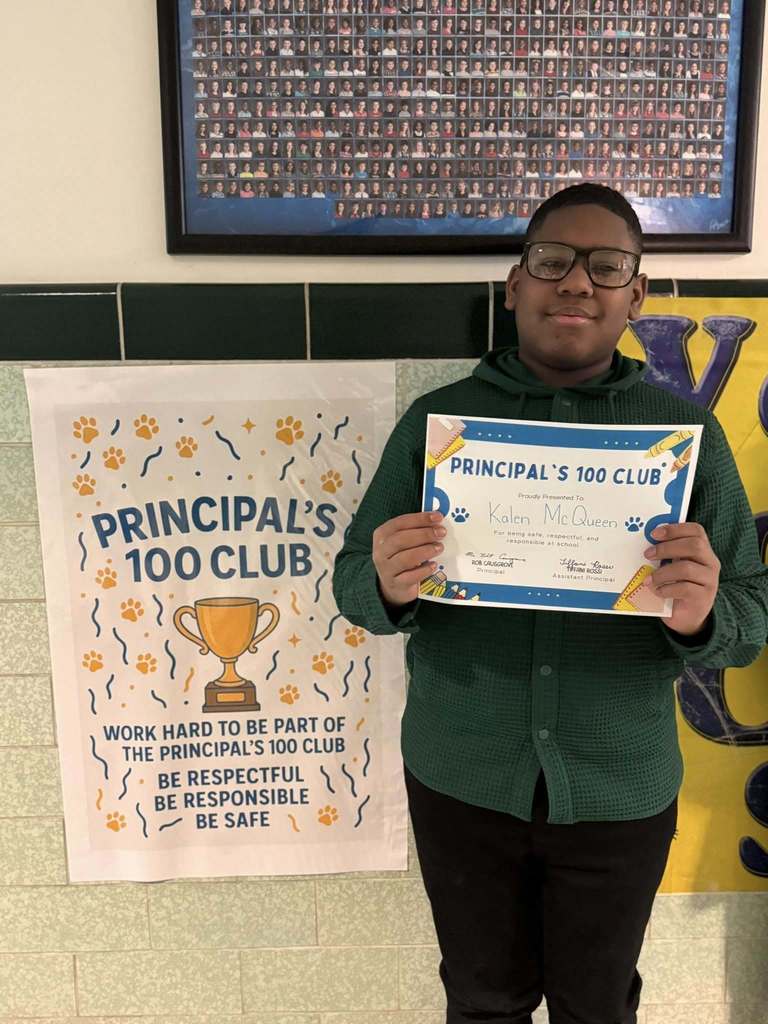 A young boy that is wearing a green button up shit is holding his Principal's 100 Club certificate. 