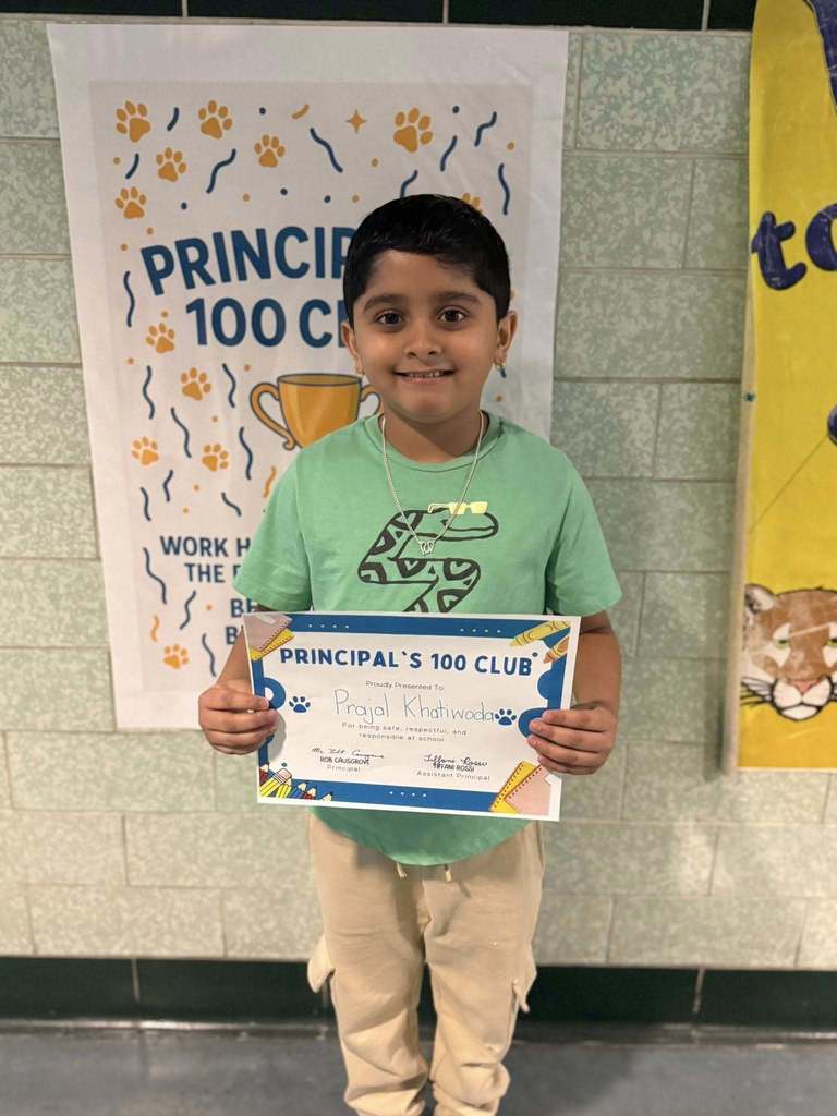 A young boy that is wearing a green shit is holding his Principal's 100 Club certificate. 
