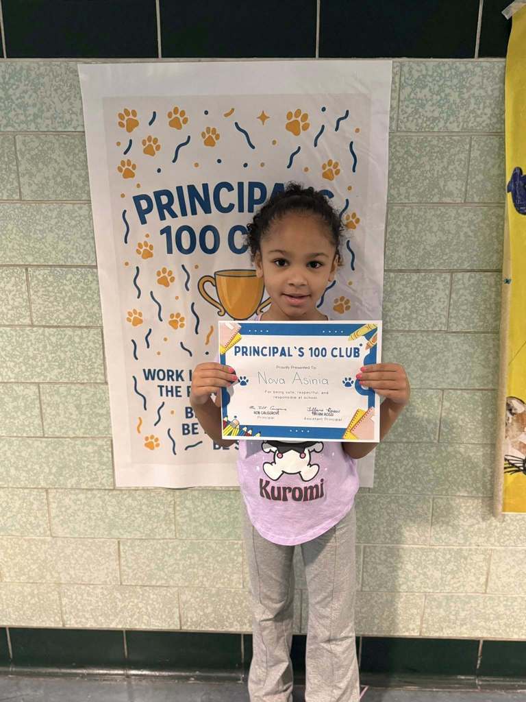 A young girl that is wearing a purple shirt and gray pants is holding his Principal's 100 Club certificate. 