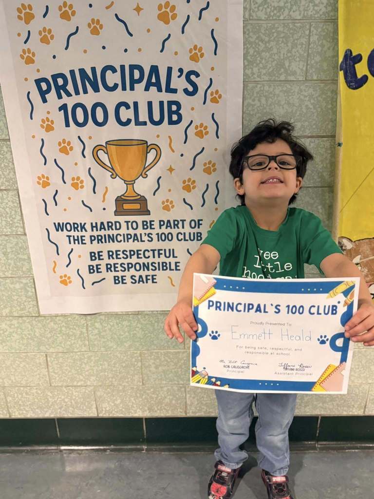 A young boy that is wearing a green shit is holding his Principal's 100 Club certificate. 