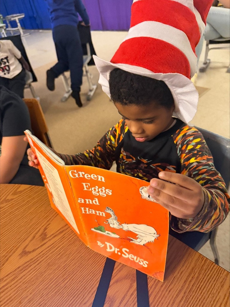 A student wearing a Dr. Seuss hat and reading the Green Eggs and Ham book. 