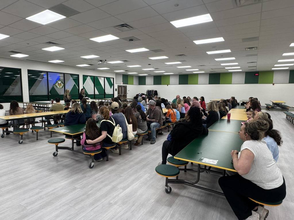A large indoor space with tables and chairs, many people seated. Tables are green. The room has white walls, a ceiling with lights, and green and white striped accents.