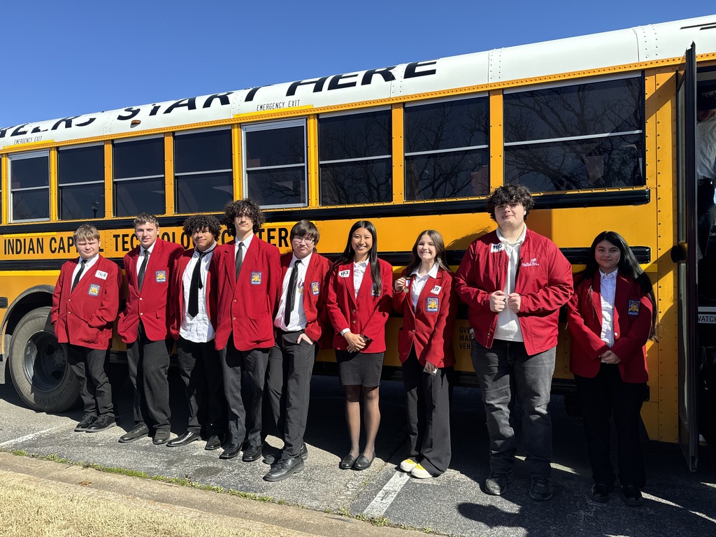 A group of people in red jackets and ties stands in front of a yellow school bus.