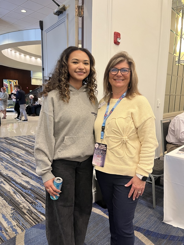 Two women in business attire stand in a modern lobby. One holds a drink, and both wear lanyards. A table and chairs are behind them.