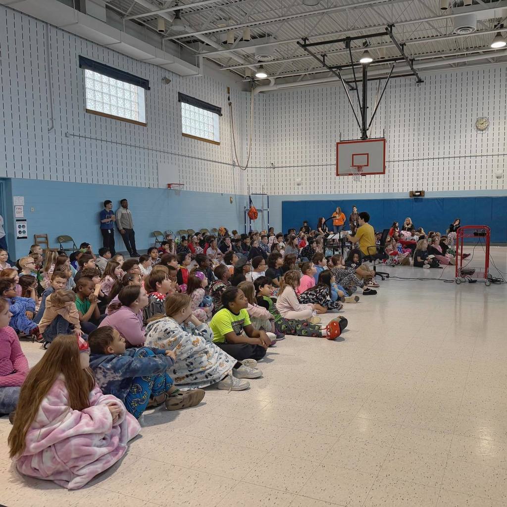Students sitting in the school gym and listening to someone read. 