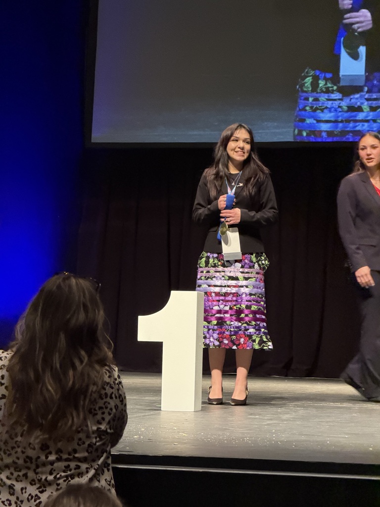 Person stands on stage next to a podium with a number one sign, holding an award.