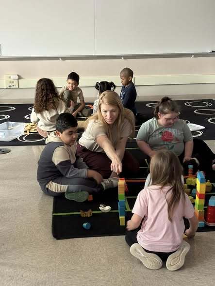 Students playing with building blocks and a teacher is pointing at one tower. 