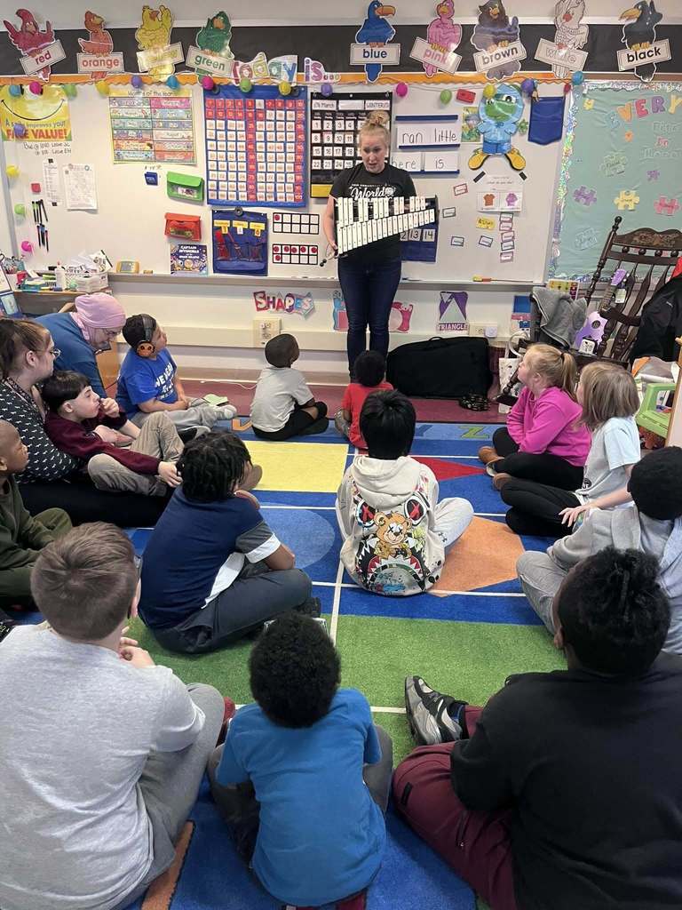 A teacher holding a xylophone and talking to students in a classroom. 