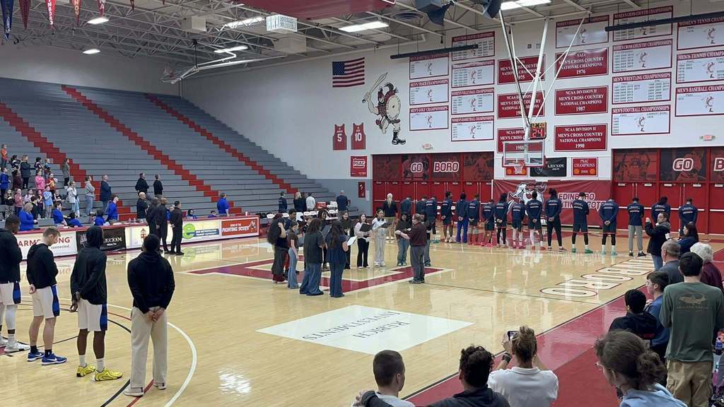 Basketball players lined up on a basketball court with students in the center singing.