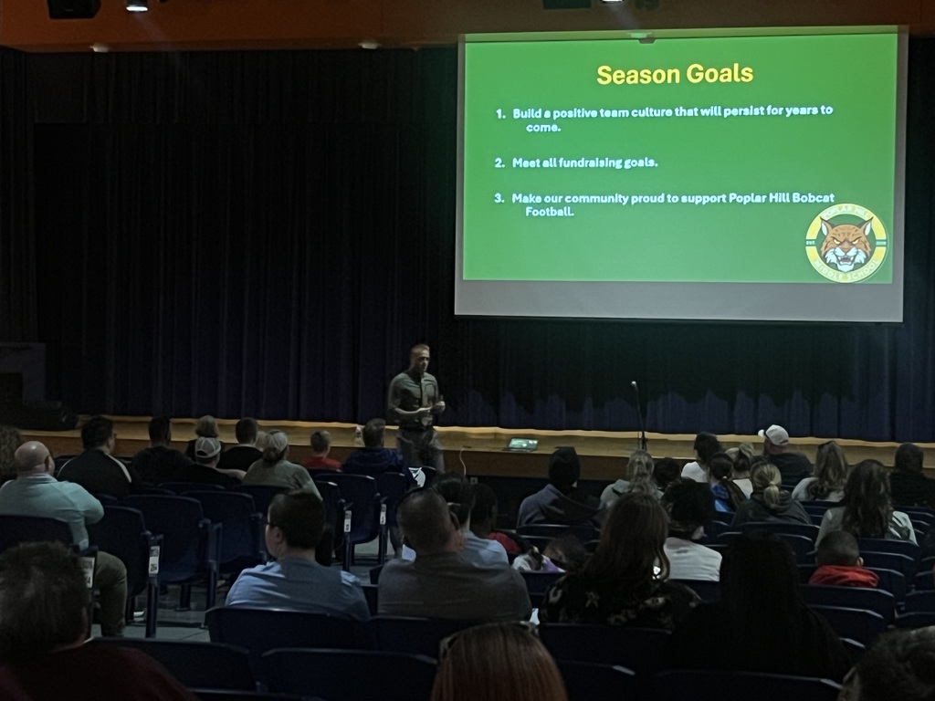 A man speaks at a podium in a classroom, facing a screen with "Season Goals" displayed. Audience members sit in rows.